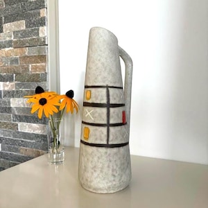 Op de afbeelding: Een hoge, getextureerde, grijze keramische kan met een handvat. De kan heeft een rasterontwerp met bruine lijnen en gele en rode accenten. Links staat een kleine glazen vaas met gele bloemen.