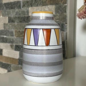 Puede incluir: Un jarrón decorativo de cerámica con base blanca y rayas horizontales grises. La parte superior presenta un diseño geométrico con triángulos naranjas, amarillos y morados. El borde es amarillo. Un elemento de decoración del hogar elegante.