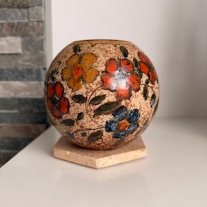 Op de afbeelding: Een ronde keramische vaas met een bloemenontwerp in oranje, rood en blauw. De vaas staat op een vierkante, lichtgeleurde stenen voet. De achtergrond is een witte muur en een stenen bakstenen muur.