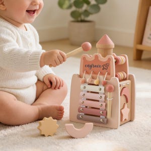 Puede incluir: Un cubo de actividades para bebés de madera rosa y natural con xilófono, ábaco y otros elementos interactivos. El cubo tiene la palabra "engrave". Incluye un mazo de madera y dos formas de madera adicionales.