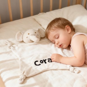 May include: A beige baby blanket with the name "Cora" embroidered in black, next to a stuffed elephant toy. The elephant is beige with striped ears. The items are on a white bed with a sleeping baby.