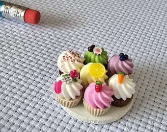 Peut inclure: Un assortiment de petits cupcakes avec des glaçages et des garnitures colorés, dont une tranche de citron, des baies et une carotte. Les cupcakes sont disposés sur une petite assiette ronde blanche. Un crayon avec une gomme rouge est en arrière-plan.