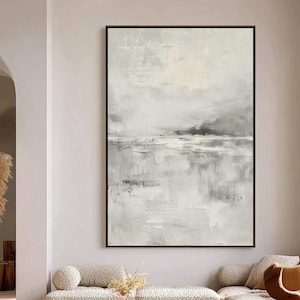 Könnte beinhalten: Ein großes, gerahmtes abstraktes Landschaftsgemälde in einem modernen Wohnzimmer. Das Kunstwerk verwendet eine gedämpfte Palette aus Grau- und Weißtönen und erzeugt eine ruhige, minimalistische Ästhetik. Der Raum umfasst einen Holztisch, ein Ecksofa und dekorative Kissen.