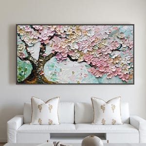 Könnte beinhalten: Gerahmtes Gemälde eines blühenden Baumes mit strukturierten rosa und cremefarbenen Blüten. Der Baumstamm ist braun, vor einem hellblauen und grünen Hintergrund. Das Kunstwerk hängt über einem weißen Sofa.