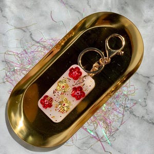 May include: A rectangular keychain with red and gold floral designs and the text "MORE YOU". The keychain has a gold-colored clasp and key ring. The keychain is on a gold-colored oval tray.