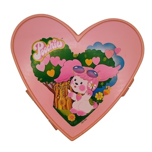 May include: A pink heart-shaped vintage Poochie toy case. The case features a cartoon dog character with pink ears and sunglasses, surrounded by a green tree with hearts. The word "Poochie" is written in a cursive font above the character.