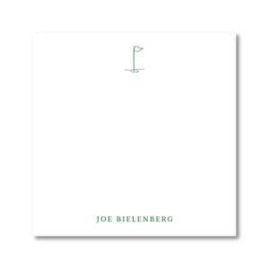 May include: A white square notepad with a minimalist green line drawing of a golf flag. The text "JOE BIELENBERG" is printed in green at the bottom of the notepad.