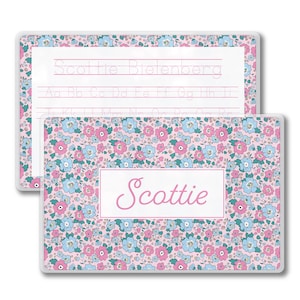 May include: Two rectangular name tags. One features the name "Scottie" in pink script on a white background, surrounded by a pink, blue, and white floral pattern. The other tag has the name "Scottie Bielenberg" and handwriting practice lines.