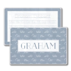May include: Two personalized placemats. The top placemat has a light blue background with handwriting practice lines and the name "Graham Lybbert". The bottom placemat has a light blue background with a pattern of construction vehicles and the name "GRAHAM".
