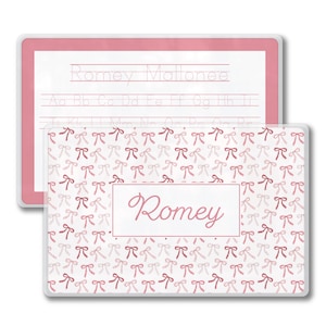 May include: Two pink and white personalized name tracing boards. The top board has the name "Romey Mallonee" and the alphabet. The bottom board has the name "Romey" in a white box, surrounded by a pattern of pink bows.