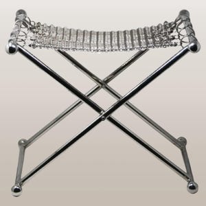 May include: A chrome-finished folding stool with a woven metal seat. The stool's legs form an X-shape, and the seat is a series of interconnected metal links. The legs end in small, rounded feet. The design is minimalist and modern.