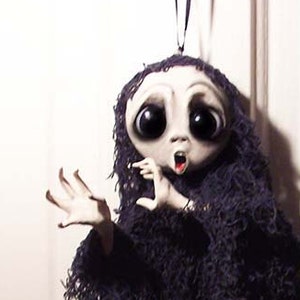 May include: A handmade ghost ornament with a white face, large eyes, and an open mouth. The ghost is wearing a black tattered fabric and is hanging from a black string.
