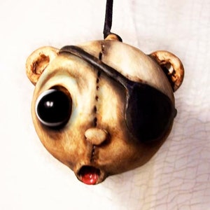 May include: A handmade, whimsical pendant in the shape of a bear's head. The bear has one large, black eye and an eye patch. The other eye is a light color. The bear's mouth is open, revealing a red tongue. The pendant is suspended from a black cord.