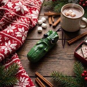 K&ouml;nnte beinhalten: Ein gr&uuml;nes, texturiertes Gew&uuml;rzgurken-Ornament mit m&uuml;rrischem Gesichtsausdruck, gro&szlig;en schwarzen Augen und einem kleinen violetten Aufh&auml;ngeband. Die Gurke liegt auf einer Holzoberfl&auml;che neben einer rot-wei&szlig;en Decke, Zimtstangen, Marshmallows und einer Tasse.