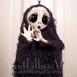 May include: A handmade doll with large eyes and a wide open mouth, wearing a black tattered fabric. The doll is hanging from a black string. The doll is a spooky Halloween decoration.