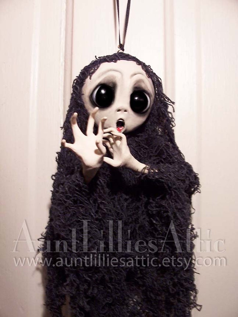 May include: A handmade doll with large eyes and a wide open mouth, wearing a black tattered fabric. The doll is hanging from a black string. The doll is a spooky Halloween decoration.