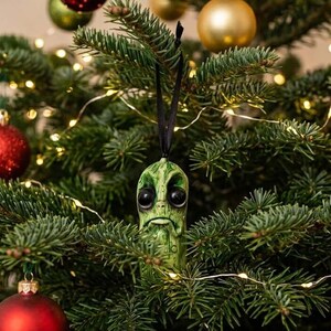 K&ouml;nnte beinhalten: Ein gr&uuml;ner, texturierter Gew&uuml;rzgurken-Anh&auml;nger mit gro&szlig;en, dunklen, gl&auml;nzenden Augen und einem nach unten gezogenen Mund, der an einem d&uuml;nnen schwarzen Band h&auml;ngt. Das Ornament hat eine detaillierte, unebene Oberfl&auml;che und h&auml;ngt an einem Tannenbaum.