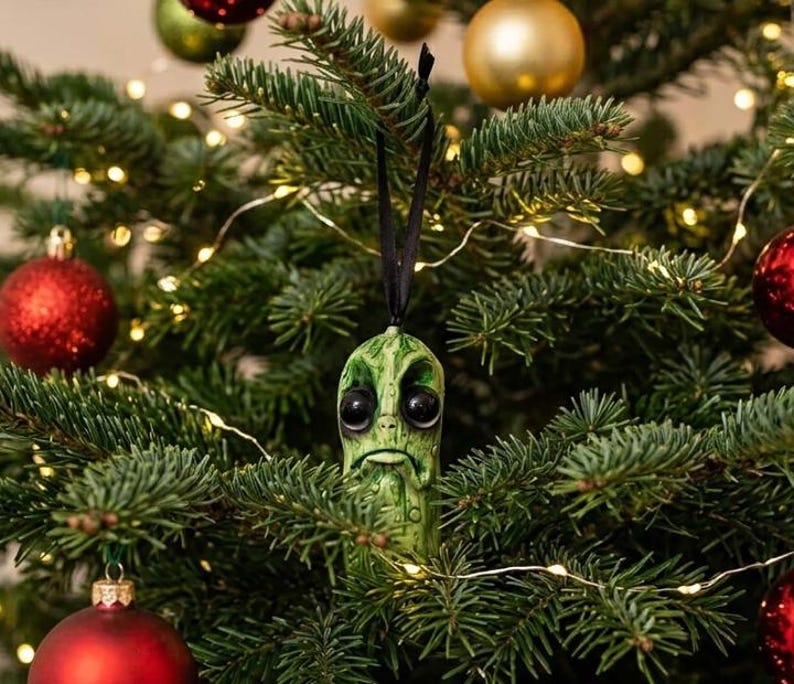 K&ouml;nnte beinhalten: Ein gr&uuml;ner, texturierter Gew&uuml;rzgurken-Anh&auml;nger mit gro&szlig;en, dunklen, gl&auml;nzenden Augen und einem nach unten gezogenen Mund, der an einem d&uuml;nnen schwarzen Band h&auml;ngt. Das Ornament hat eine detaillierte, unebene Oberfl&auml;che und h&auml;ngt an einem Tannenbaum.