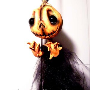 May include: A handmade Halloween decoration featuring a pumpkin-faced doll with stitched details, wearing a black tulle skirt. The doll has large, black eyes and a stitched mouth. The doll is hanging from a black string.