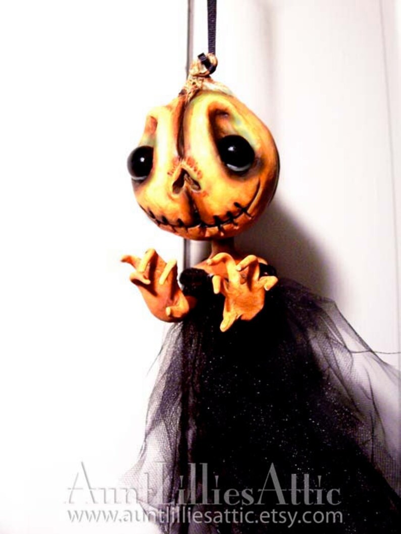 May include: A handmade Halloween decoration featuring a pumpkin-faced doll with stitched details, wearing a black tulle skirt. The doll has large, black eyes and a stitched mouth. The doll is hanging from a black string.