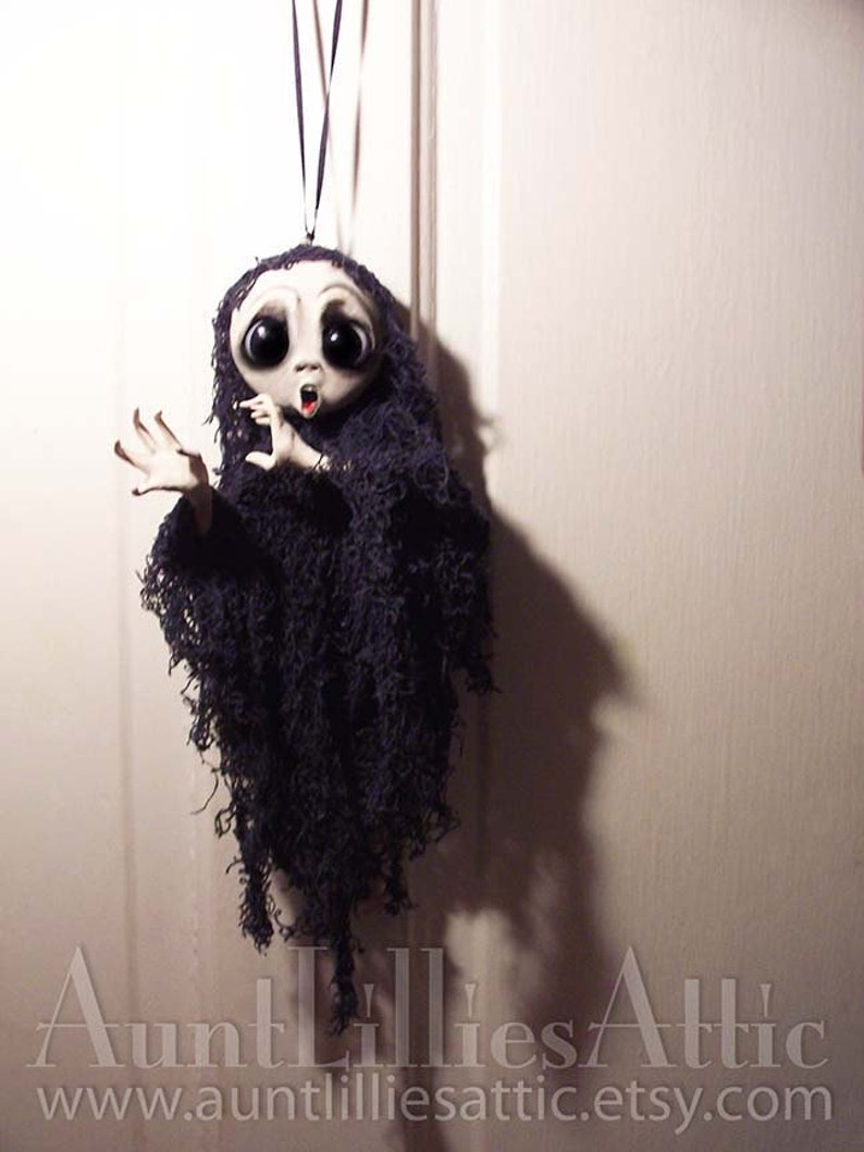 May include: A handmade ghost ornament with a white face, large eyes, and an open mouth. The ghost is wearing a black tattered fabric and is hanging from a black string.