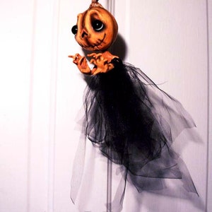 May include: A handmade pumpkin-shaped doll with a black tulle skirt. The doll has painted eyes and a stitched mouth. The doll is hanging from a white door.
