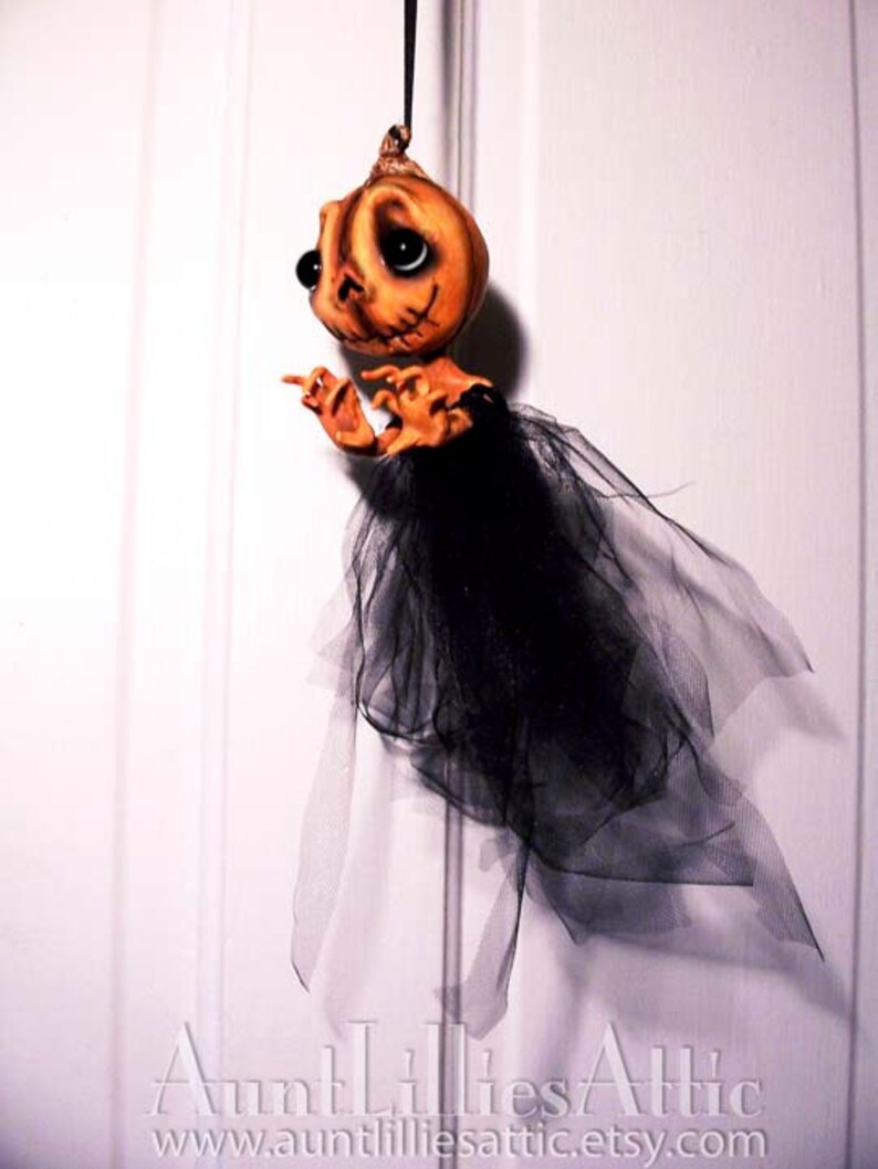 May include: A handmade pumpkin-shaped doll with a black tulle skirt. The doll has painted eyes and a stitched mouth. The doll is hanging from a white door.