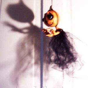 May include: A hanging Halloween decoration featuring a pumpkin-shaped doll with a black tulle skirt and a shadow of the doll on a white wall. The doll has a painted face with large eyes and a stitched mouth.