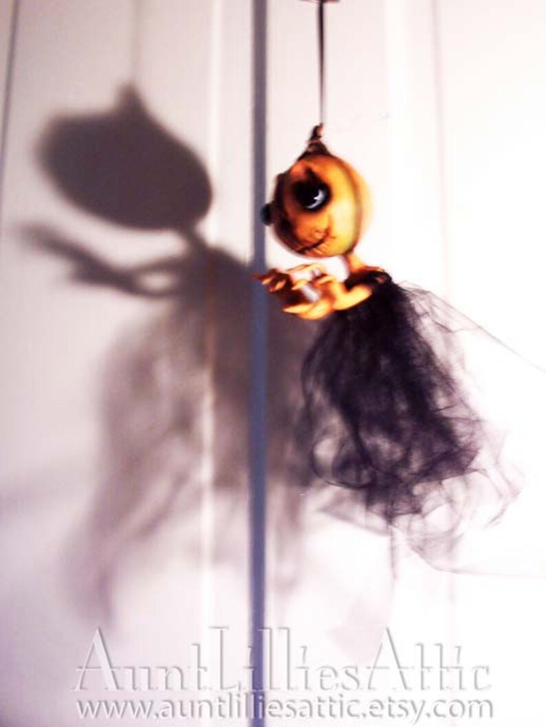 May include: A hanging Halloween decoration featuring a pumpkin-shaped doll with a black tulle skirt and a shadow of the doll on a white wall. The doll has a painted face with large eyes and a stitched mouth.