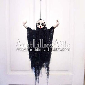 May include: A black and white ghost doll with a long, tattered black gown hanging from a white door. The doll has a white face with black eyes and a red mouth.