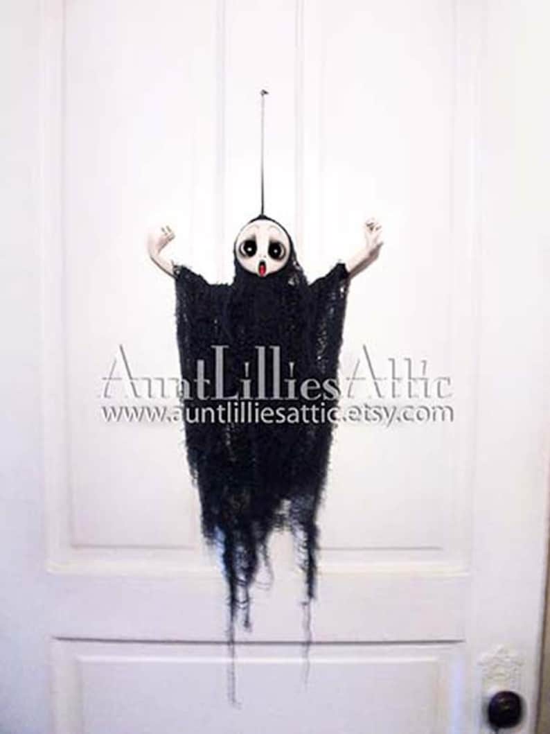 May include: A black and white ghost doll with a long, tattered black gown hanging from a white door. The doll has a white face with black eyes and a red mouth.