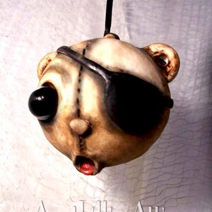 May include: A unique, handmade pendant featuring a stylized bear head with an eye patch. The bear has one large black eye and a stitched design. The mouth is open with a red interior. The pendant is suspended from a black cord. The text "Aunt Lillies Attic" is at the bottom.