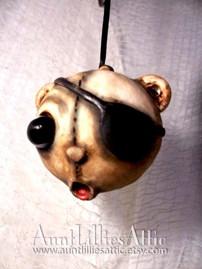 May include: A unique, handmade pendant featuring a stylized bear head with an eye patch. The bear has one large black eye and a stitched design. The mouth is open with a red interior. The pendant is suspended from a black cord. The text "Aunt Lillies Attic" is at the bottom.