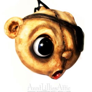 May include: A handcrafted, whimsical mask with a large, expressive eye and a stitched appearance. The mask is primarily tan with dark brown accents, an eye patch, and a red mouth. The mask has a unique, artistic design.