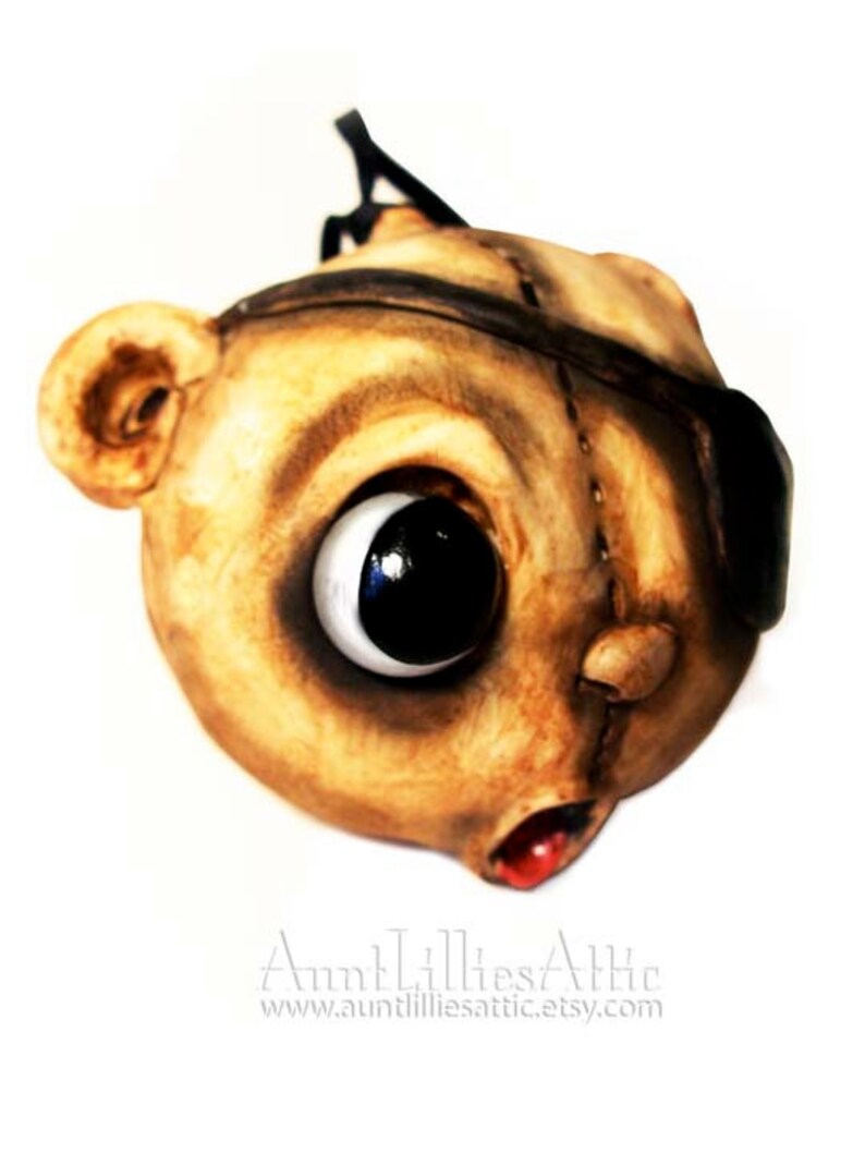 May include: A handcrafted, whimsical mask with a large, expressive eye and a stitched appearance. The mask is primarily tan with dark brown accents, an eye patch, and a red mouth. The mask has a unique, artistic design.