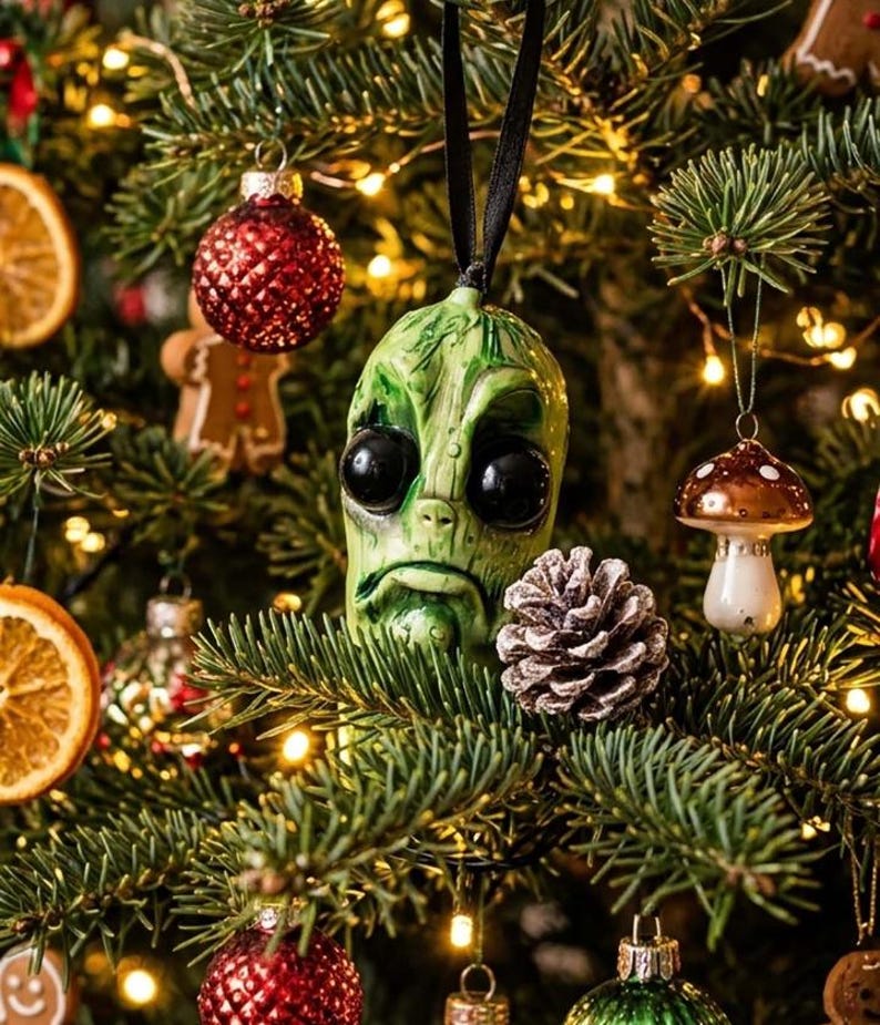 K&ouml;nnte beinhalten: Ein gr&uuml;ner, au&szlig;erirdisch geformter Weihnachtsbaumanh&auml;nger mit gro&szlig;en schwarzen Augen und einem herabh&auml;ngenden Mund, aufgeh&auml;ngt an einem schwarzen Band. Das Ornament hat eine texturierte, verwitterte Oberfl&auml;che und h&auml;ngt zwischen Tannenzweigen.