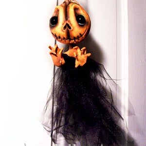 May include: A handmade Halloween decoration featuring a pumpkin-faced doll with black eyes and a black tulle skirt. The doll is hanging from a string and has a distressed, vintage look.