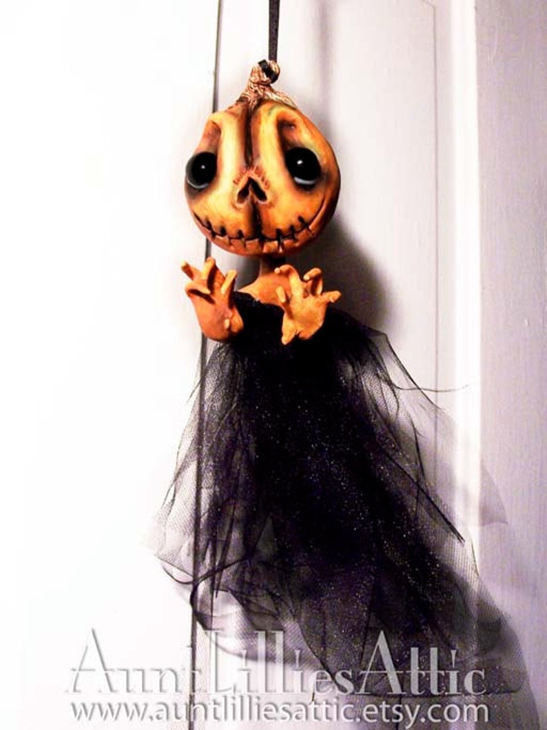 May include: A handmade Halloween decoration featuring a pumpkin-faced doll with black eyes and a black tulle skirt. The doll is hanging from a string and has a distressed, vintage look.