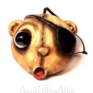 May include: A unique, handmade mask in the shape of a bear's head. The mask is painted in shades of tan and brown, with one large black eye and an eye patch. The mouth is open, revealing a red interior. A black cord is attached for hanging.