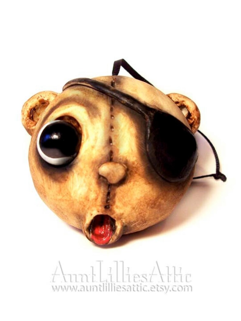 May include: A unique, handmade mask in the shape of a bear's head. The mask is painted in shades of tan and brown, with one large black eye and an eye patch. The mouth is open, revealing a red interior. A black cord is attached for hanging.
