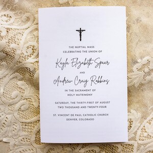 May include: A white wedding program with black text and a cross icon. The program lists the names Kayla Elizabeth Spaar and Andrew Craig Robbins, along with the date and location of the ceremony. The program is on a cream lace surface.