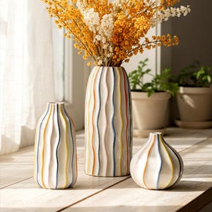 Op de afbeelding: Drie keramische vazen met verticale golvende strepen in verschillende kleuren. De hoogste vaas bevat een boeket van gedroogde gele en witte bloemen. De vazen staan op een houten oppervlak bij een raam.