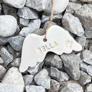 K&ouml;nnte beinhalten: Ein wei&szlig;es Keramik-Engelsfl&uuml;gel-Ornament mit dem Namen "BELLA" und einem Pfotenabdruck. Das Ornament h&auml;ngt an einer nat&uuml;rlichen Schnur und liegt auf einem Bett aus grauen und wei&szlig;en Steinen.