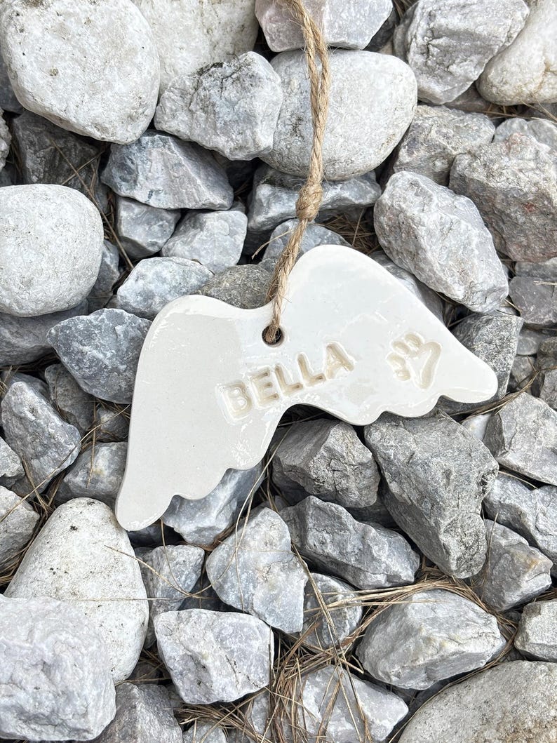 K&ouml;nnte beinhalten: Ein wei&szlig;es Keramik-Engelsfl&uuml;gel-Ornament mit dem Namen "BELLA" und einem Pfotenabdruck. Das Ornament h&auml;ngt an einer nat&uuml;rlichen Schnur und liegt auf einem Bett aus grauen und wei&szlig;en Steinen.