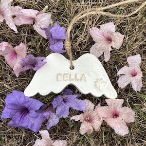 K&ouml;nnte beinhalten: Wei&szlig;es Keramik-Engelsfl&uuml;gel-Ornament mit dem Namen "BELLA" und einem Pfotenabdruck. Das Ornament h&auml;ngt an einer nat&uuml;rlichen Schnur und ist von rosa und lila Blumen auf trockenem Gras umgeben.