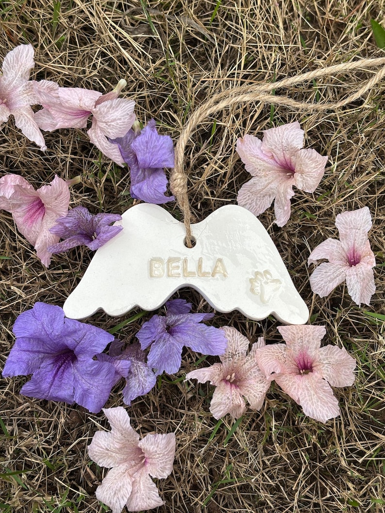 K&ouml;nnte beinhalten: Wei&szlig;es Keramik-Engelsfl&uuml;gel-Ornament mit dem Namen "BELLA" und einem Pfotenabdruck. Das Ornament h&auml;ngt an einer nat&uuml;rlichen Schnur und ist von rosa und lila Blumen auf trockenem Gras umgeben.