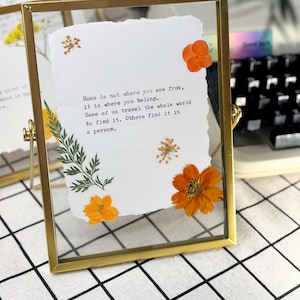 May include: A gold-framed glass picture frame with a white paper insert. The paper features pressed orange flowers and green foliage, along with the quote: "Home is not where you are from, it is where you belong."