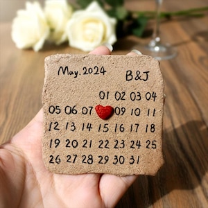 May include: A handmade, square, textured, tan-coloured calendar for May 2024, with a red heart marking a date. The calendar is inscribed with the initials "B&J" and the days of the month. The calendar is held in a hand.