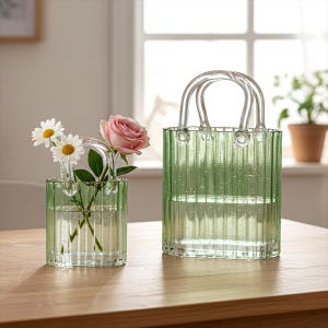 May include: Two green glass vases shaped like handbags, one holding daisies and a pink rose. The vases have clear handles and vertical ribbed detailing. They sit on a wooden surface, with a window and plants in the background.