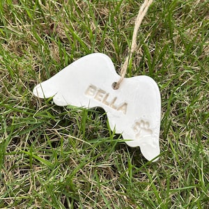 K&ouml;nnte beinhalten: Ein wei&szlig;er Keramik-Engelsfl&uuml;gel-Ornament mit dem Namen "BELLA" und einem Pfotenabdruck. Das Ornament h&auml;ngt an einer Schnur aus Naturfaser vor dem Hintergrund von gr&uuml;nem Gras.